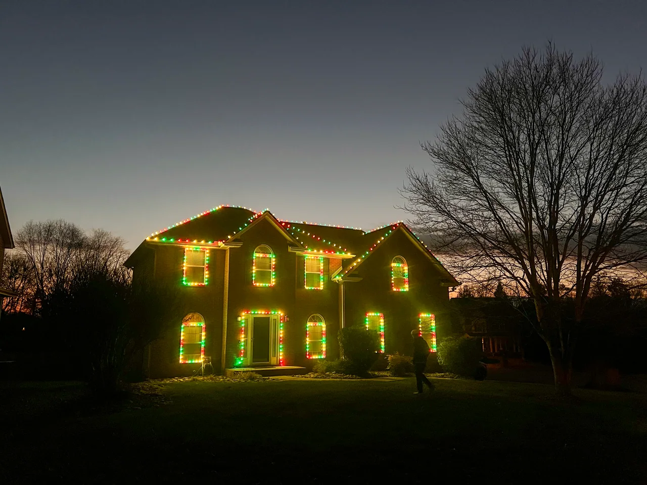 Red, green and warm lights outlining a brick home’s roofline and windows at dusk