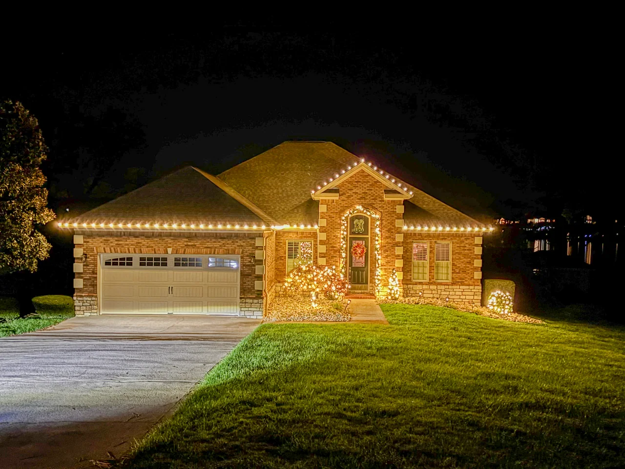 Single-story brick home with warm-white roofline lights and illuminated shrubs at night