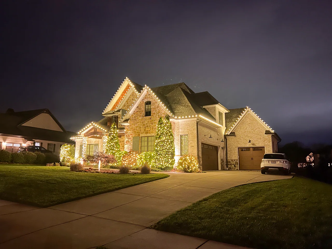 Stone home with warm-white Christmas roofline lights and wrapped evergreens, viewed from the driveway