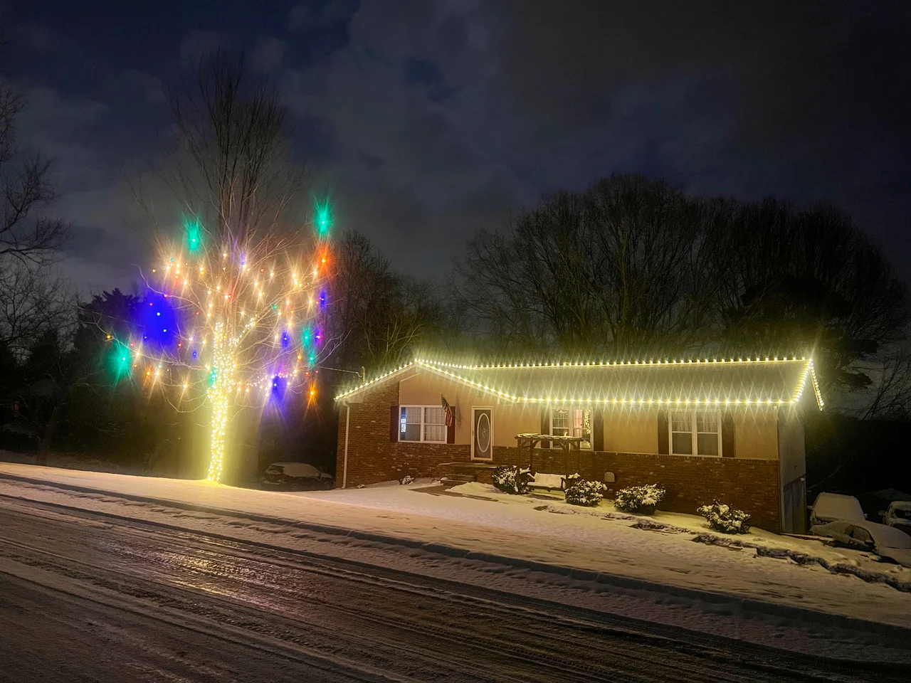 Warm-white roofline lights and a colorful illuminated tree outside a brick home in the snow
