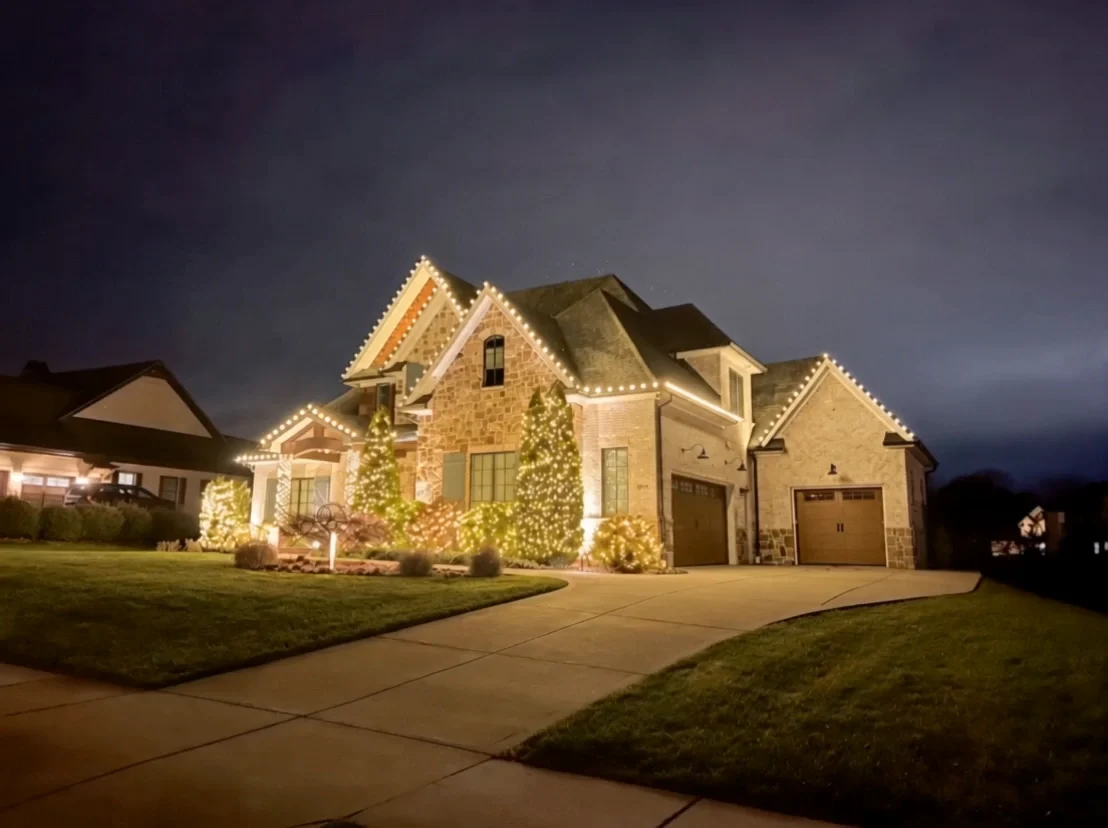 A stone home glowing with warm-white Christmas roofline and landscape lights